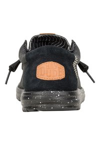 Zapato negro de ante tipo slip-on con un patrón texturizado y un parche de cuero marrón. Presenta una suela negra con manchas y cordones elásticos.