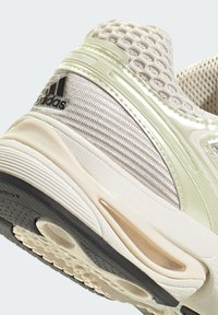 Svetlozafarbená športová obuv s priedušným zvrškom, textúrovanými striebornými akcentmi, gumovou podrážkou a charakteristickým čiernym logom Adidas. Mierne zakrivený dizajn.