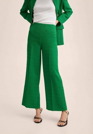 Žena v zelených kalhotách s vysokým pasem a širokými nohavicemi, ladícím blejzru, bílém topu a černých sandálech se pásky, stojící s rukama v kapsách.