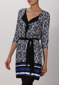 Czarna i biała sukienka w geometryczny wzór z rękawami do trzech czwartych, czarnym paskiem w talii i niebieskimi poziomymi paskami u dołu.