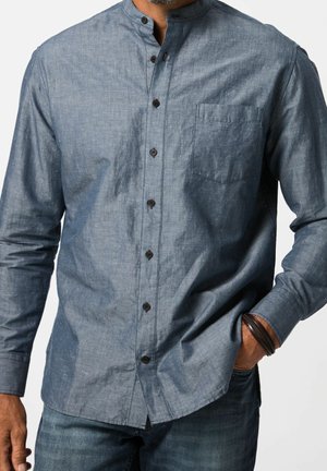 Man wearing a blue chambray long-sleeve shirt with a mandarin collar, black buttons, chest pocket, and jeans, hand in pocket with leather bracelet.
