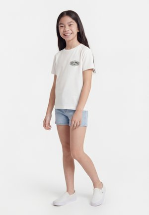 Fille souriante aux longs cheveux foncés, vêtue d'un t-shirt blanc avec un motif graphique, un short en jean bleu clair et des baskets blanches à enfiler.
