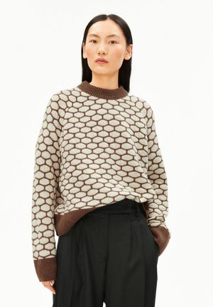 Frau trägt einen braun-beigen Pullover mit sechseckigem Muster und schwarze, hoch taillierte Hosen, steht mit einer Hand in der Tasche.