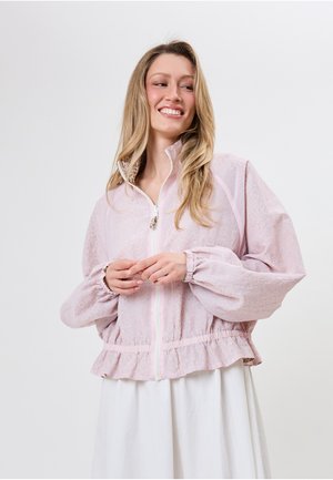 Lachende vrouw met lang blond haar die een lichtroze jas met ritssluiting draagt met pofmouwen en een gerimpelde zoom, gecombineerd met een witte rok.