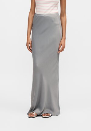 Γυναίκα που φοράει μακριά, λεία γκρι φούστα με λευκό ριγωτό αμάνικο τοπ και λευκά σανδάλια, στέκεται μπροστά σε απλό φόντο.