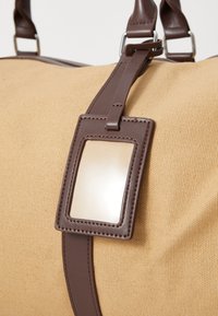 Sac de sport tan avec des accents en cuir marron. Présente une étiquette rectangulaire lisse pour l'identification. Surface en tissu texturé.