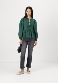 Blusa peplum em tecido verde escuro com decote redondo e detalhe em laço, combinada com jeans cinzentos e sapatilhas creme, segurando uma mala preta.