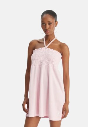 Vrouw draagt lichtroze, strapless, geplooide minijurk met halterneck en grote oorringen, staand tegen een witte achtergrond.