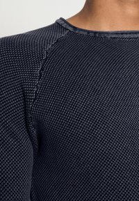 Tessuto testurizzato blu navy con motivo a diamante, caratterizzato da un collo a giro e dettagli di cuciture visibili lungo le cuciture delle spalle.