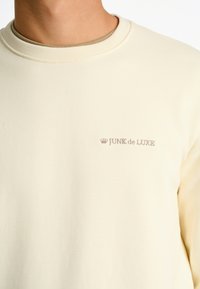Felpa color crema con colletto a coste e logo ricamato "JUNK de LUXE" sul petto. Texture morbida e liscia.