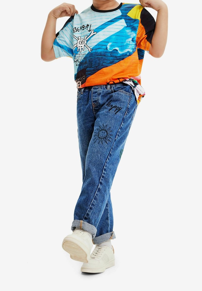 Farverig grafisk t-shirt med blå, orange og gule mønstre; parret med lyseblå denimjeans med sol- og "glad" tryk. Hvide sneakers.