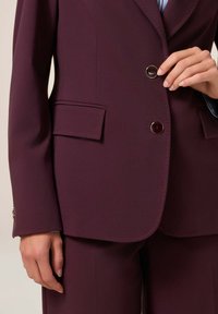 Primo piano di una persona che allaccia una giacca su misura di colore viola scuro con tasche a pattina e pantaloni abbinati.