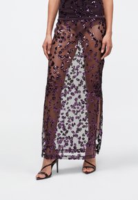 Femme portant une jupe noire transparente ornée de paillettes violettes et noires sur une lingerie noire, associée à des sandales à talons hauts noires.