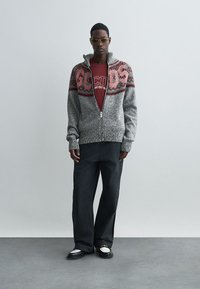 Grijze gebreide ritstrui met een rood geometrisch patroon, gedragen over een wijnrode shirt, in combinatie met zwarte brede broek en zwarte schoenen.