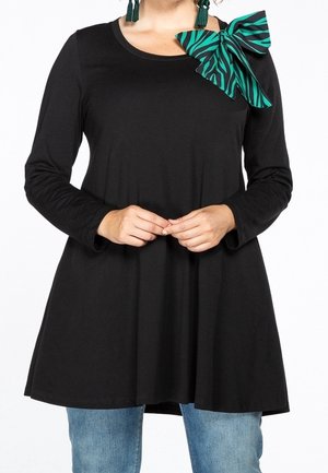 Femme portant une tunique noire à manches longues avec un grand nœud rayé vert et noir sur l'épaule gauche, associée à un jean bleu.