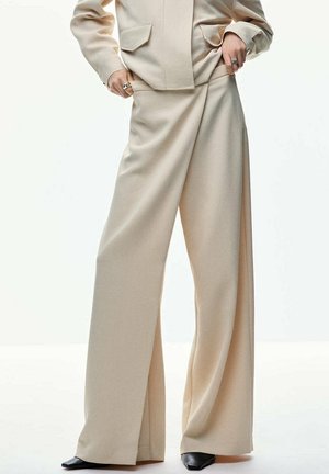 Persona che indossa pantaloni beige a gamba larga con un dettaglio a incrocio, abbinati a una giacca beige coordinata e scarpe nere a punta.