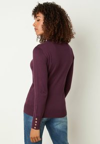 Lila Pullover mit langen Ärmeln und taillierter Passform, gerippte Bündchen mit goldenen Knöpfen und glatter Textur. Tragen mit blauen Jeans.