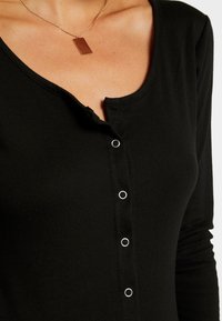 Haut noir à manches longues en tissu côtelé avec un décolleté arrondi, doté de fermetures à boutons et de accents en métal argenté le long de l'avant.