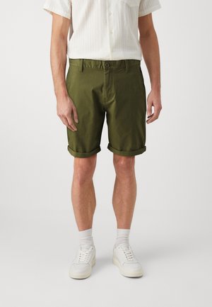 Mand iført olivengrønne opklappede shorts, hvid kortærmet skjorte med knapper, hvide sokker og hvide sneakers, står op af en hvid baggrund.