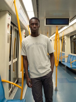 Jonge man in licht T-shirt en donkere broek staat in een lege metrowagon met blauwe stoelen en gele handgrepen.