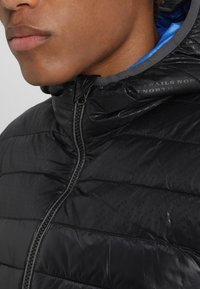 Svart pufferjacka med dragkedja fram, quiltad textur och en grå krage. Insidan har en blå foder som visar subtil branding.