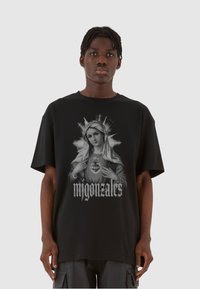Sort bomulds T-shirt med et grafisk print af en kvinde med en glorie og teksten "migonazales" nedenfor. Løs pasform, korte ærmer.