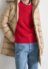 Person klädd i en beige puffjacka över en röd kabelstickad tröja och ljusblå jeans, som står framför en enfärgad vit bakgrund.