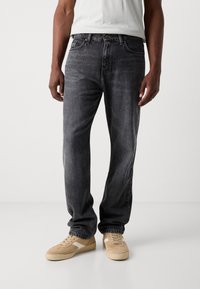 Jeans de mezclilla gris oscuro con un corte relajado, cinco bolsillos y cierre de botón, combinados con zapatillas beige que tienen franjas blancas.