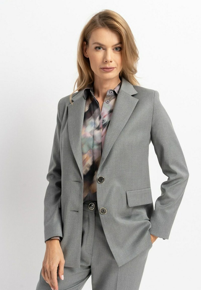 Grauer maßgeschneiderter Blazer mit zwei Knöpfen, Reverskragen und Taschen, getragen über einer mehrfarbigen gemusterten Bluse. Weicher, strukturierter Stoff.