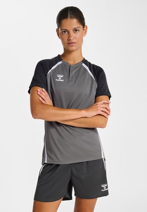 Frau steht mit verschränkten Armen, trägt graue und schwarze Hummel-Sportbekleidung, bestehend aus einem kurzärmeligen Shirt und Shorts.