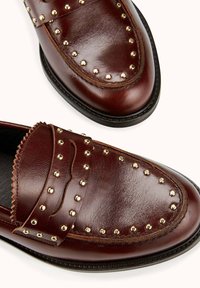 Mocassins en cuir marron ornés de clous dorés ronds le long des bords, avec un design festonné sur la bride et une texture lisse.