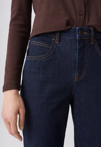 Persoon draagt donkerblauwe spijkerbroek met oranje stiksel en een bruin langemouwen top, hand ontspannen langs het lichaam.