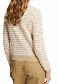 Vrouw met lang golvend haar, gekleed in een beige en witte gestructureerde lange mouwen trui en lichtbruine broek, gezien van achteren.