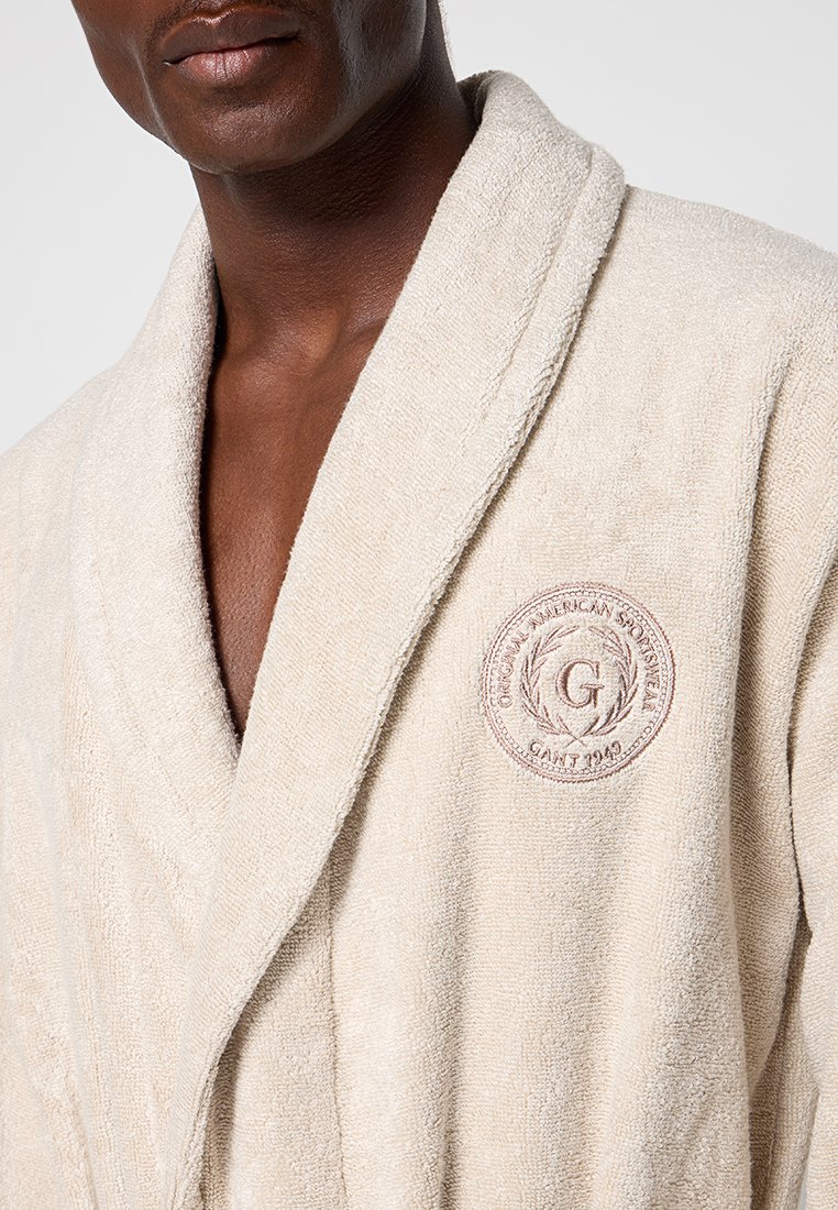 Ciemnoskóry mężczyzna ubrany w kremowy, teksturowany szlafrok z okrągłym haftowanym logo na piersi, na którym widnieje napis "GANT 1949."