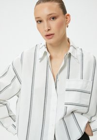 White button-up shirt featuring gray vertical stripes, with a patch pocket and a collared neckline. Made from lightweight fabric.
