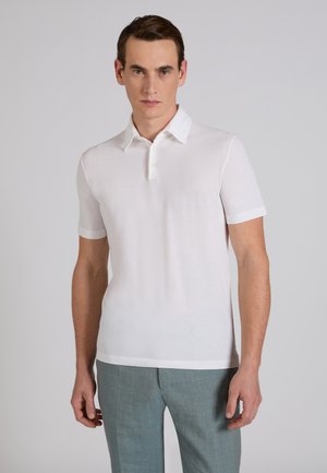 Młody mężczyzna ubrany w białą koszulkę polo z krótkim rękawem i jasnoszare spodnie, stojący na tle jednolitego jasnoszarego tła.