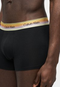 Ανδρικός κορμός που φοράει μαύρα εσώρουχα boxer Calvin Klein με ζώνη μέσης σε γραντίντ κίτρινο χρώμα και το λογότυπο Calvin Klein.