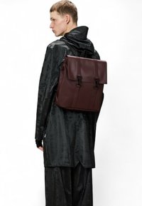 Zaino in pelle marrone con patta, due spallacci e una superficie liscia. Lo sfondo è bianco e il modello indossa un cappotto nero e lucido.