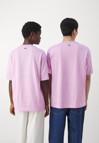 T-shirt di cotone rosa con maniche corte, caratterizzate da un piccolo logo sulla schiena. Indossate con jeans blu e pantaloni bianchi.