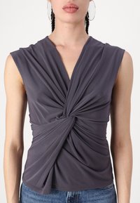 Femme portant un top gris foncé à nœud tordu sans manches avec un décolleté en V profond et un jean bleu, vu des épaules à la taille.