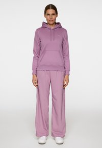 Felpa con cappuccio di un leggero colore viola con tasca frontale abbinata a pantaloni larghi dello stesso colore. Entrambi gli articoli hanno una consistenza liscia.