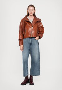 Giubbotto in pelle marrone corto con colletto ampio, abbinato a jeans blu a gamba larga e stivaletti voluminosi bordeaux. Sotto, una semplice maglietta grigia.