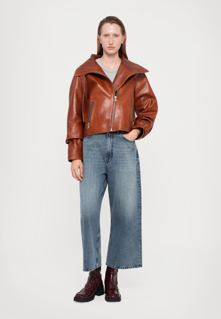 Veste en cuir marron courte avec un large col, associée à un jean large bleu et des bottes chunky bordeaux. Simple haut gris en dessous.