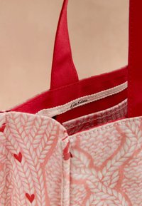 Punane kangast ostukott, millel on tekstureeritud roosa lehemuster ja väikesed punased südamed. Varustatud tugeva punaste õlarihmadega ja sees oleva kootud sildiga.