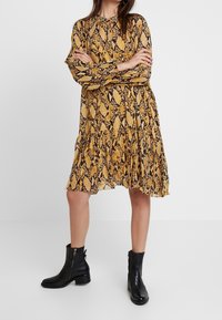 Šaty s potiskem hadí kůže v žluté a černé barvě, dlouhé rukávy, kravata na krku a vrstvený spodní lem; doplněné černými kotníkovými botami.