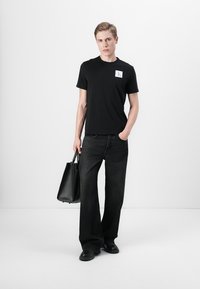 T-shirt noir à manches courtes avec un patch logo, associé à un jean large sombre et des chaussures noires, portant un sac fourre-tout noir. Design minimaliste.