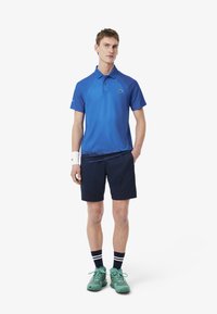 Blå pikétröja med gradientdesign, med krage och logotyp, paret med marinblå shorts och gröna träningsskor med blå detaljer.