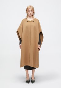 Καπέλο από καμήλα με αβέλτιστη εφαρμογή, με χρυσή αλυσίδα στο λαιμό. Κάτω από αυτό, φαίνεται ένα σκούρο, πλεκτό φόρεμα με ρίγες.