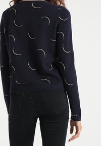 Granatowy sweter z wzorami półksiężyca wykonanymi z metalicznej nici, ściągacze na mankietach i dole, dopasowany fason, noszony na czarnych dżinsach.