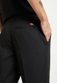 Pantaloni neri con una texture liscia, dotati di tasche posteriori e di un piccolo logo Dickies. La vita è posizionata più in alto, enfatizzando la vestibilità.